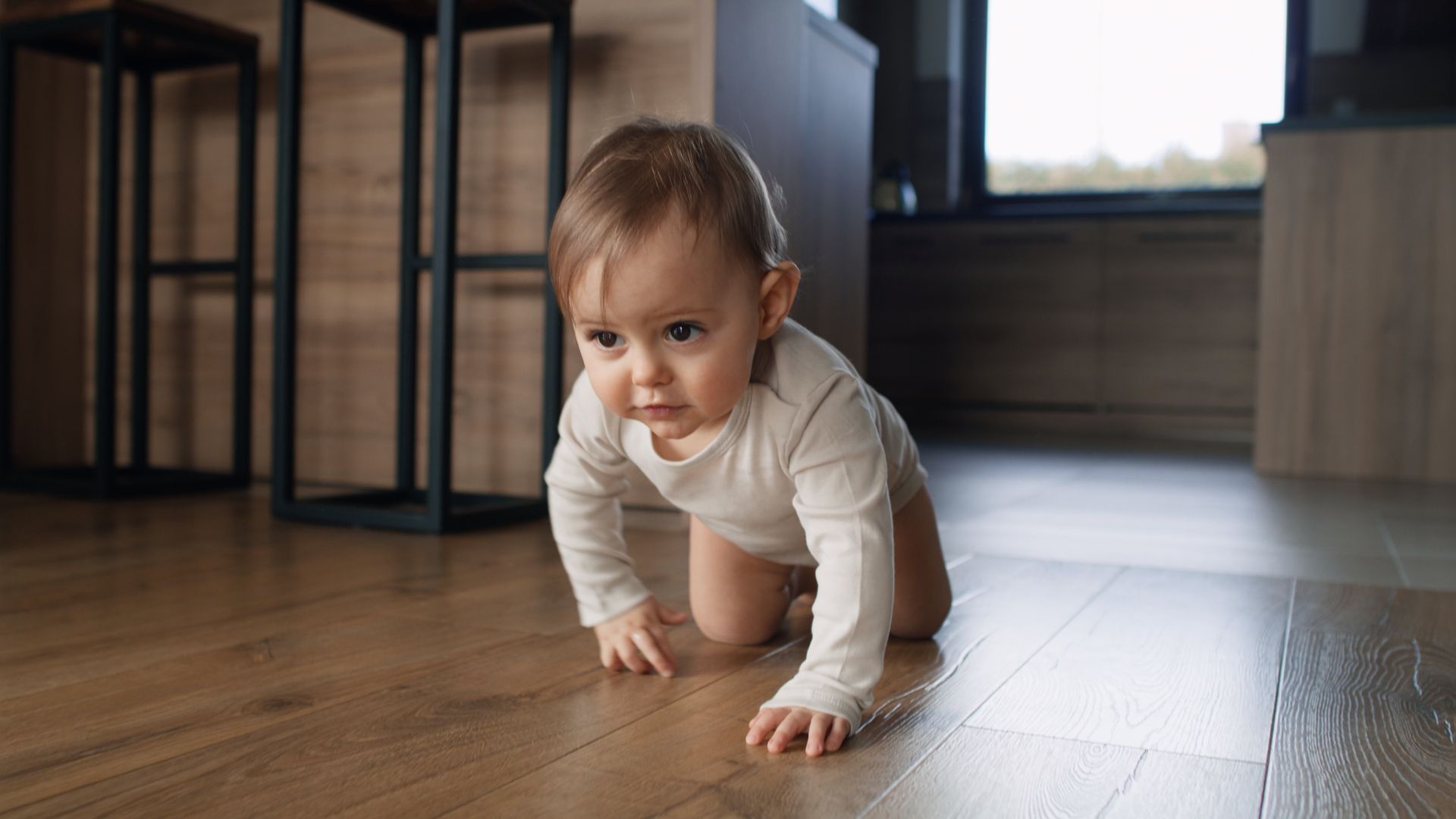 Baby crawling on a wooden floor in a room with a window and furniture in the background